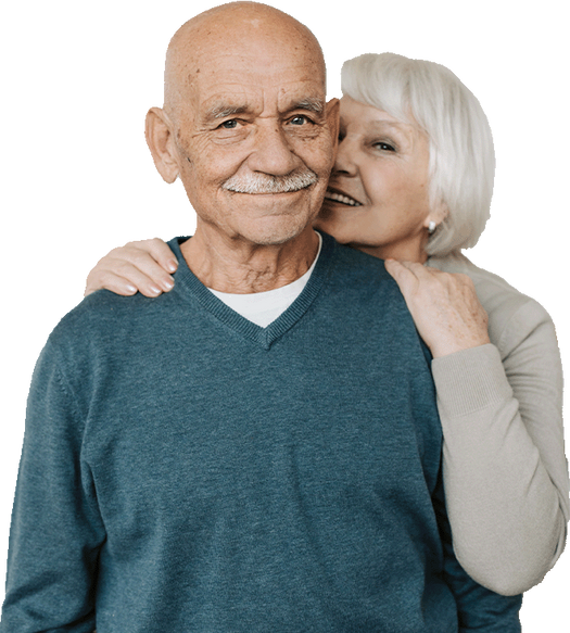 Senior couple smiling at the camera, the woman has her arms around the mans shoulders.